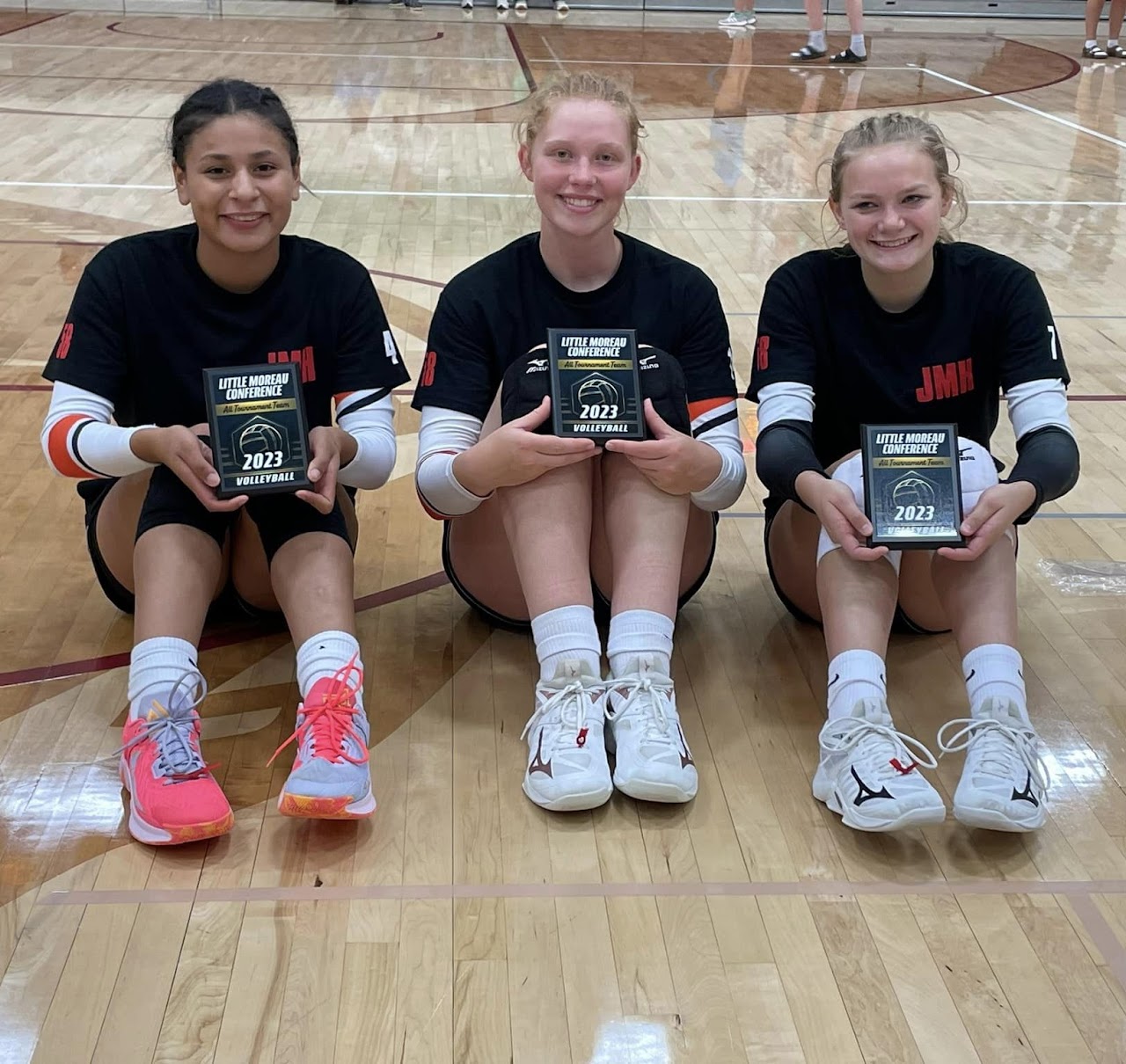 Three players sitting on the gym floor holding their All-Tourney team awards in front of them